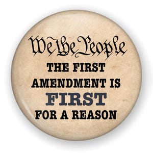 May include: A round pin-back button with a beige, parchment-like background. The text reads "We the People" in a black script font, followed by "THE FIRST AMENDMENT IS FIRST FOR A REASON" in bold, black, block-style capital letters.