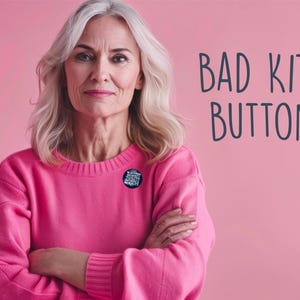 May include: A woman with gray hair wearing a pink sweater, arms crossed, with a button pinned on the sweater. The background is pink, and the text "BAD KITTY BUTTONS" is on the right side of the image.