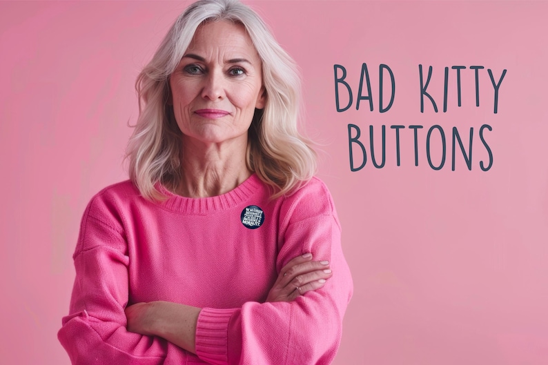 May include: A woman with gray hair wearing a pink sweater, arms crossed, with a button pinned on the sweater. The background is pink, and the text "BAD KITTY BUTTONS" is on the right side of the image.