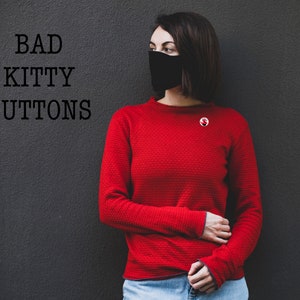May include: A woman wearing a red long-sleeved shirt with a black face mask and a red pin with a white handprint. The text "BAD KITTY BUTTONS" is written in black on a gray wall behind her.
