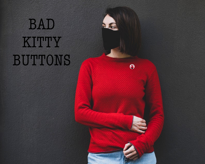 May include: A woman wearing a red long-sleeved shirt with a black face mask and a red pin with a white handprint. The text "BAD KITTY BUTTONS" is written in black on a gray wall behind her.