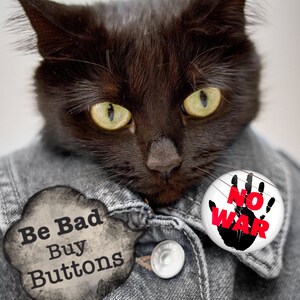 May include: A black cat wearing a denim jacket with two buttons. One button says "Be Bad Buy Buttons" and the other says "No War" with a black handprint and red text.