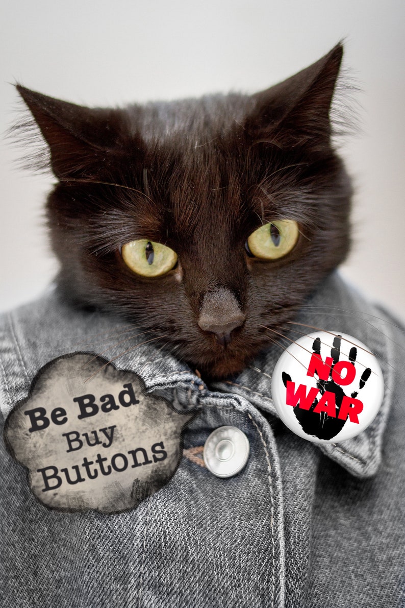 May include: A black cat wearing a denim jacket with two buttons. One button says "Be Bad Buy Buttons" and the other says "No War" with a black handprint and red text.