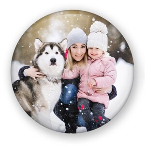May include: A woman and a young girl are smiling and holding a gray and white dog in a snowy winter scene. The woman is wearing a gray beanie and a blue jacket. The girl is wearing a white beanie and a pink jacket. The dog is standing on its hind legs.