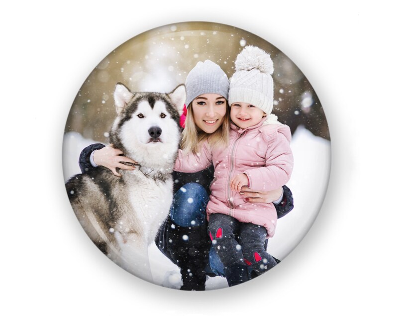 May include: A woman and a young girl are smiling and holding a gray and white dog in a snowy winter scene. The woman is wearing a gray beanie and a blue jacket. The girl is wearing a white beanie and a pink jacket. The dog is standing on its hind legs.