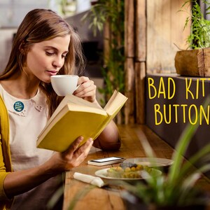 K&ouml;nnte beinhalten: Eine Person liest ein Buch und trinkt Kaffee. Sie tr&auml;gt eine gelbe Strickjacke &uuml;ber einem wei&szlig;en Hemd mit einem angesteckten Knopf. Der Text "BAD KITTY BUTTONS" ist gelb geschrieben.