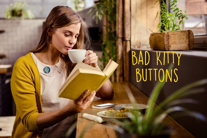 K&ouml;nnte beinhalten: Eine Person liest ein Buch und trinkt Kaffee. Sie tr&auml;gt eine gelbe Strickjacke &uuml;ber einem wei&szlig;en Hemd mit einem angesteckten Knopf. Der Text "BAD KITTY BUTTONS" ist gelb geschrieben.