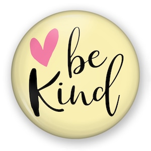May include: A round, light yellow button with the words "be kind" in black script. A pink heart is above the word "kind". The button has a glossy finish and a white background.