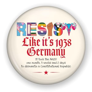 May include: A round, off-white button with the word "RESIST" in colorful letters. Below, in red gothic script, it reads "Like it's 1938 Germany." The text "It took the NAZIS one month, 3 weeks and 2 days to dismantle a Constitutional Republic" is also included.