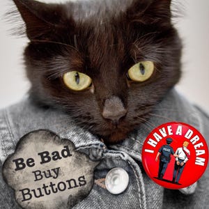 May include: A black cat with yellow eyes wearing a denim jacket and two buttons. One button reads "Be Bad Buy Buttons" and the other says "I Have A Dream" with a graphic of a police officer and a person in handcuffs.