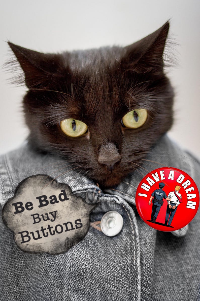 May include: A black cat with yellow eyes wearing a denim jacket and two buttons. One button reads "Be Bad Buy Buttons" and the other says "I Have A Dream" with a graphic of a police officer and a person in handcuffs.