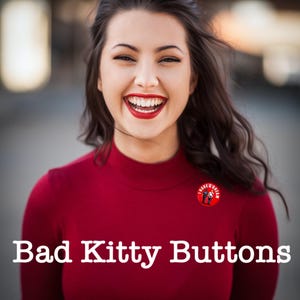 May include: A person wearing a red turtleneck top with the text "Bad Kitty Buttons" in white. A red button with the text "I Have A Dream" is pinned on the top. The person is smiling with red lipstick.