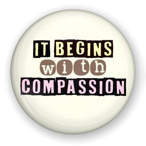 May include: A white button with black text that reads "IT BEGINS with COMPASSION". The words "with" are in brown and the word "COMPASSION" is in pink.