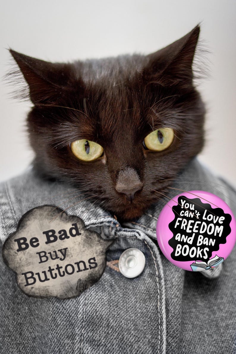Puede incluir: Un gato negro con una chaqueta vaquera con dos botones. El gato tiene ojos amarillos y lleva dos botones. Un bot&oacute;n dice "Be Bad Buy Buttons" y el otro dice "You can't love freedom and ban books."