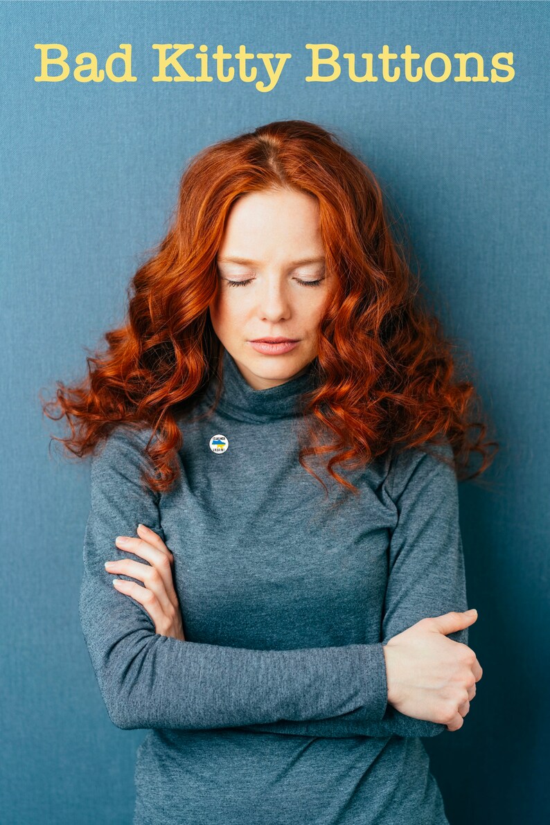 May include: A woman with long, curly red hair is wearing a gray turtleneck sweater with a small blue and yellow button. She has her arms crossed and is looking down.