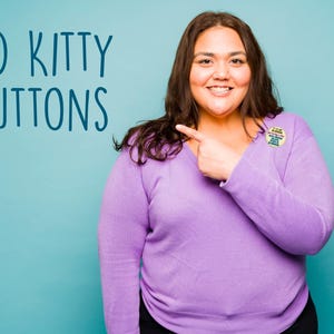Puede incluir: Una persona con un su&eacute;ter morado de manga larga y pantalones negros, se&ntilde;alando un bot&oacute;n en su pecho. El bot&oacute;n tiene texto. Las palabras "BAD KITTY BUTTONS" est&aacute;n escritas en azul sobre un fondo turquesa.