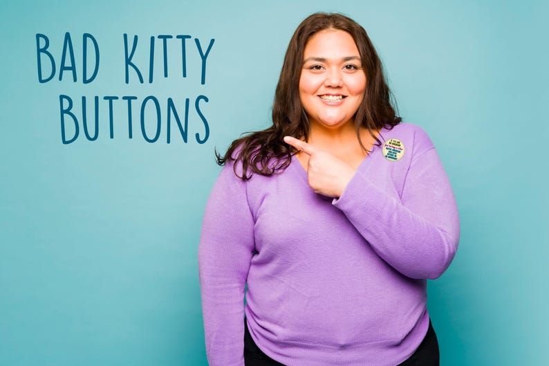 Puede incluir: Una persona con un su&eacute;ter morado de manga larga y pantalones negros, se&ntilde;alando un bot&oacute;n en su pecho. El bot&oacute;n tiene texto. Las palabras "BAD KITTY BUTTONS" est&aacute;n escritas en azul sobre un fondo turquesa.