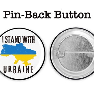 May include: A white button with a black border and the text "I Stand With Ukraine" in black. The button features a blue and yellow outline of the country of Ukraine. The back of the button is silver with a pin.