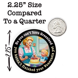 Op de afbeelding: Een ronde button van ongeveer 5,7 cm diameter met een retro illustratie van een vrouw en kind die een taart versieren. De rand van de button bevat de tekst: "No you can't have Greenland yet, you haven't finished your Venezuela yet."