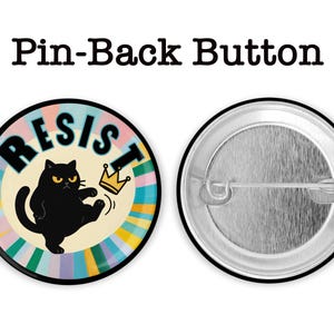May include: A round pin-back button with a black cat kicking a gold crown. The word "RESIST" is written in black letters on a colorful background. The button's back is silver.