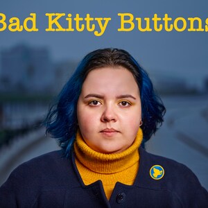 Pu&ograve; includere: Una donna con i capelli blu indossa una giacca blu scuro sopra un maglione a collo alto giallo. Indossa uno spillo blu e giallo con una colomba. Il testo "Bad Kitty Buttons" &egrave; in cima all'immagine.