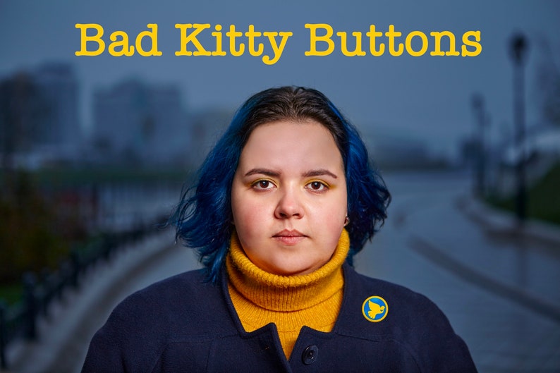 Pu&ograve; includere: Una donna con i capelli blu indossa una giacca blu scuro sopra un maglione a collo alto giallo. Indossa uno spillo blu e giallo con una colomba. Il testo "Bad Kitty Buttons" &egrave; in cima all'immagine.