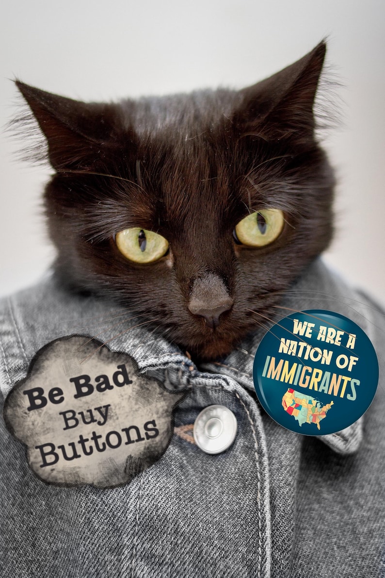 K&ouml;nnte beinhalten: Eine schwarze Katze tr&auml;gt eine Jeansjacke mit zwei Kn&ouml;pfen. Die Katze hat gelbe Augen. Ein Knopf sagt "Be Bad Buy Buttons" und der andere sagt "We Are A Nation of Immigrants" mit einer Karte der USA.