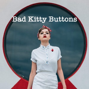 May include: A woman wearing a white collared shirt with a red hand print pin, a white skirt, and a red and white headband. The text "Bad Kitty Buttons" is above her head.