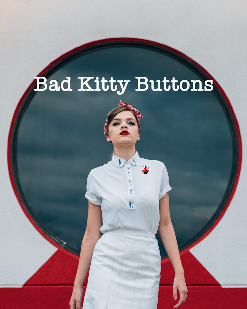 May include: A woman wearing a white collared shirt with a red hand print pin, a white skirt, and a red and white headband. The text "Bad Kitty Buttons" is above her head.