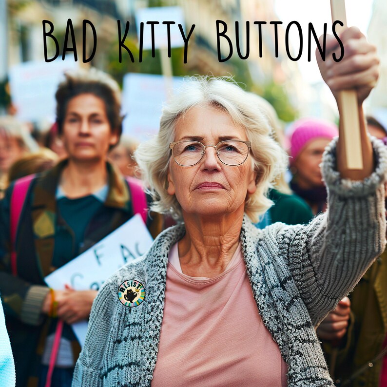 May include: Image of a protest with the text "BAD KITTY BUTTONS" at the top. A woman with glasses and a "RESIST" button on her sweater raises a sign. Other people are in the background, some holding signs.