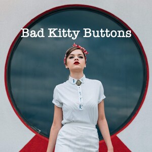 K&ouml;nnte beinhalten: Eine Frau tr&auml;gt ein wei&szlig;es Hemd mit Kragen und einem schwarzen Knopf mit goldenen Details, ein rot-wei&szlig;es Stirnband und einen wei&szlig;en Rock. Der Text "Bad Kitty Buttons" steht &uuml;ber ihrem Kopf.