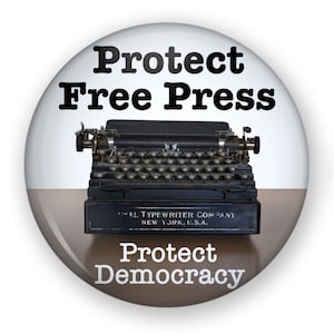 May include: A white button with black text that reads "Protect Free Press" and "Protect Democracy". A vintage typewriter is pictured in the center of the button.
