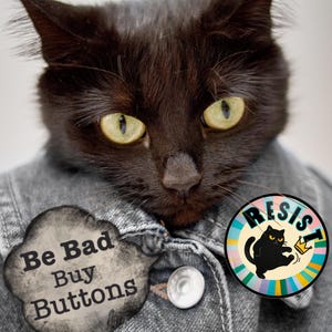 May include: A black cat with yellow eyes wearing a denim jacket. The cat has two buttons on the jacket and two buttons. A cloud-shaped button reads "Be Bad Buy Buttons." A round button reads "RESIST" with a cat wearing a crown.