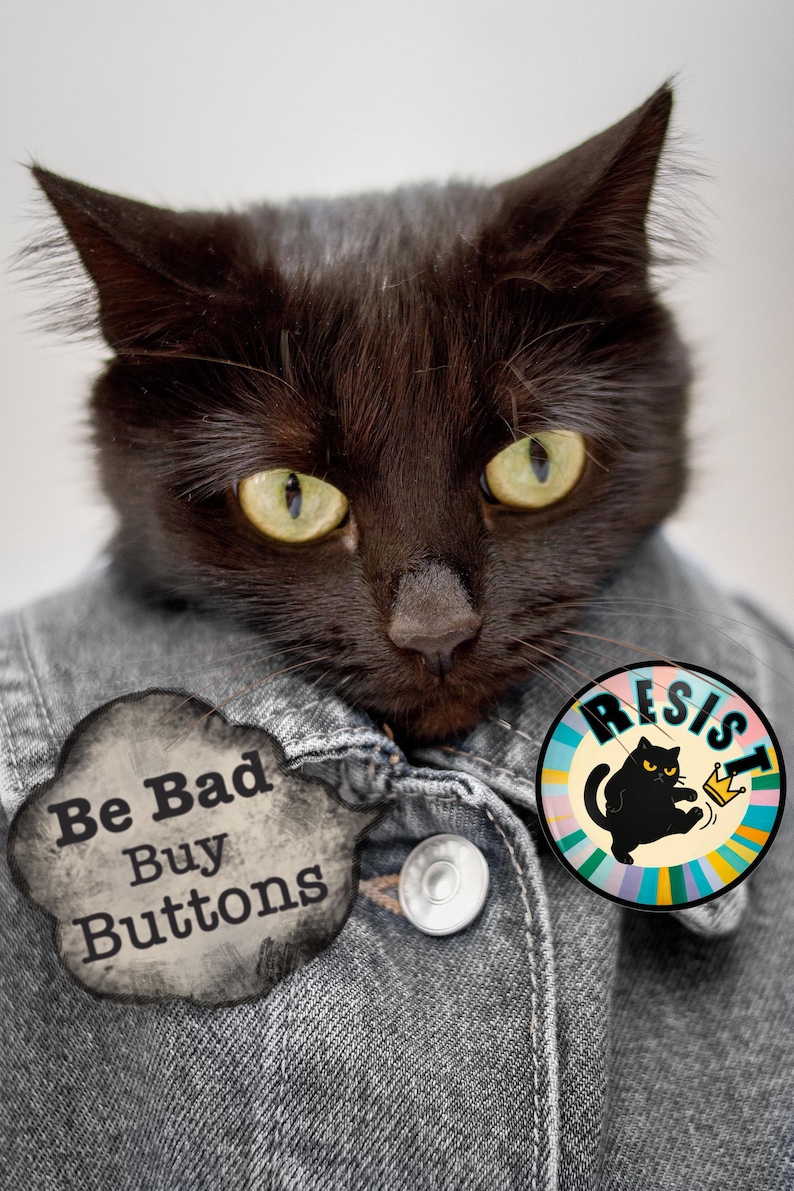 May include: A black cat with yellow eyes wearing a denim jacket. The cat has two buttons on the jacket and two buttons. A cloud-shaped button reads "Be Bad Buy Buttons." A round button reads "RESIST" with a cat wearing a crown.