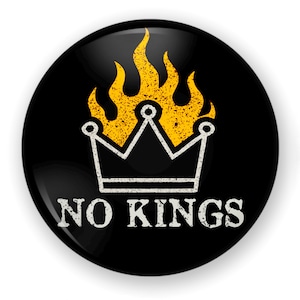 May include: A black round button with a white crown design and yellow flames. The text "NO KINGS" is printed in white below the crown. The button has a glossy finish.