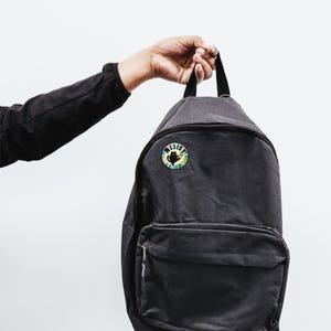 May include: A black backpack with a round button featuring a black cat silhouette and the word "RESIST" in a colorful design. The text "BAD Kitty BUTTONS" is above the backpack.