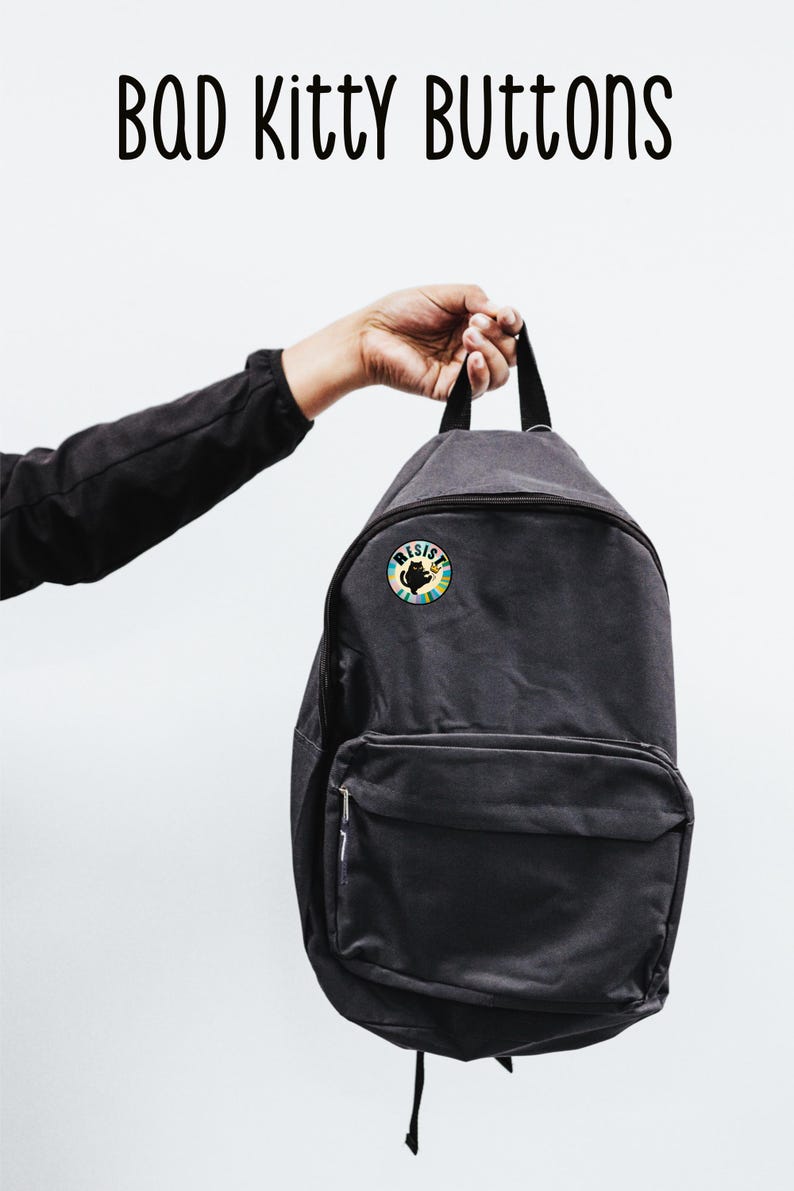 May include: A black backpack with a round button featuring a black cat silhouette and the word "RESIST" in a colorful design. The text "BAD Kitty BUTTONS" is above the backpack.