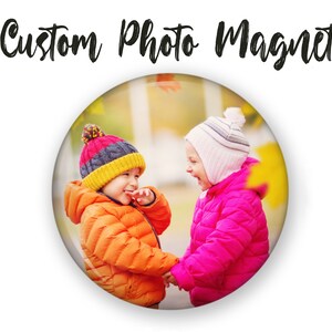 May include: Two children wearing winter coats and hats are smiling at each other in a park setting. The child on the left is wearing an orange coat and a multi-colored hat. The child on the right is wearing a pink coat and a white hat.