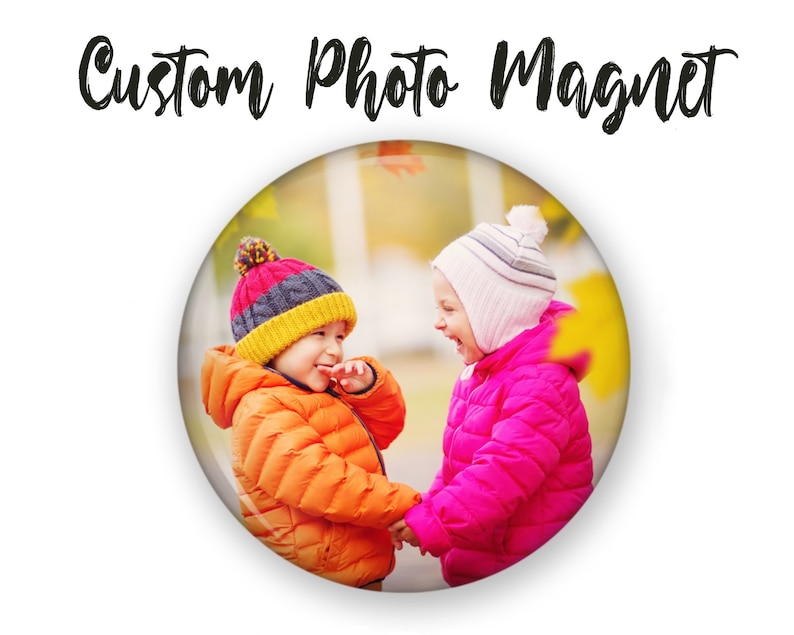 May include: Two children wearing winter coats and hats are smiling at each other in a park setting. The child on the left is wearing an orange coat and a multi-colored hat. The child on the right is wearing a pink coat and a white hat.