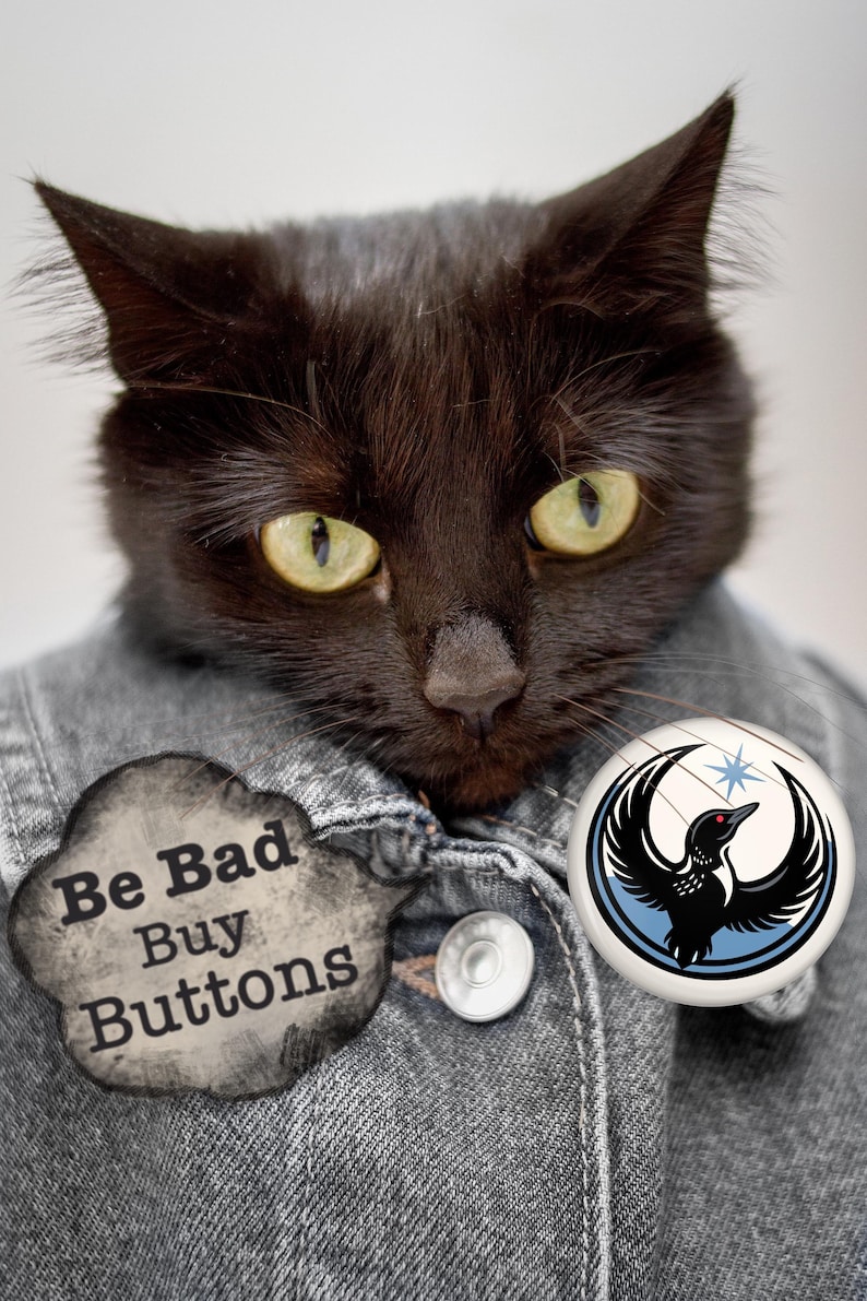 May include: A black cat wearing a denim jacket with two buttons. The cat has yellow eyes and is wearing two buttons. One button says "Be Bad Buy Buttons" and the other has a bird design.
