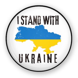 May include: A white button with a black border featuring a blue and yellow outline of Ukraine with the text "I Stand With Ukraine" in black.