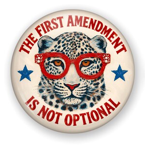 May include: A circular pinback button featuring a leopard wearing bright red glasses. The text THE FIRST AMENDMENT IS NOT OPTIONAL is printed in red around the leopard. Two blue stars are positioned on the left and right sides of the leopard.