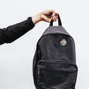 Op de afbeelding: Een donkergrijze rugzak met een patch met een cartoonafbeelding. De rugzak heeft een voorvak en een handvat aan de bovenkant. De tekst "BAD Kitty BUTTONS" staat boven de rugzak.