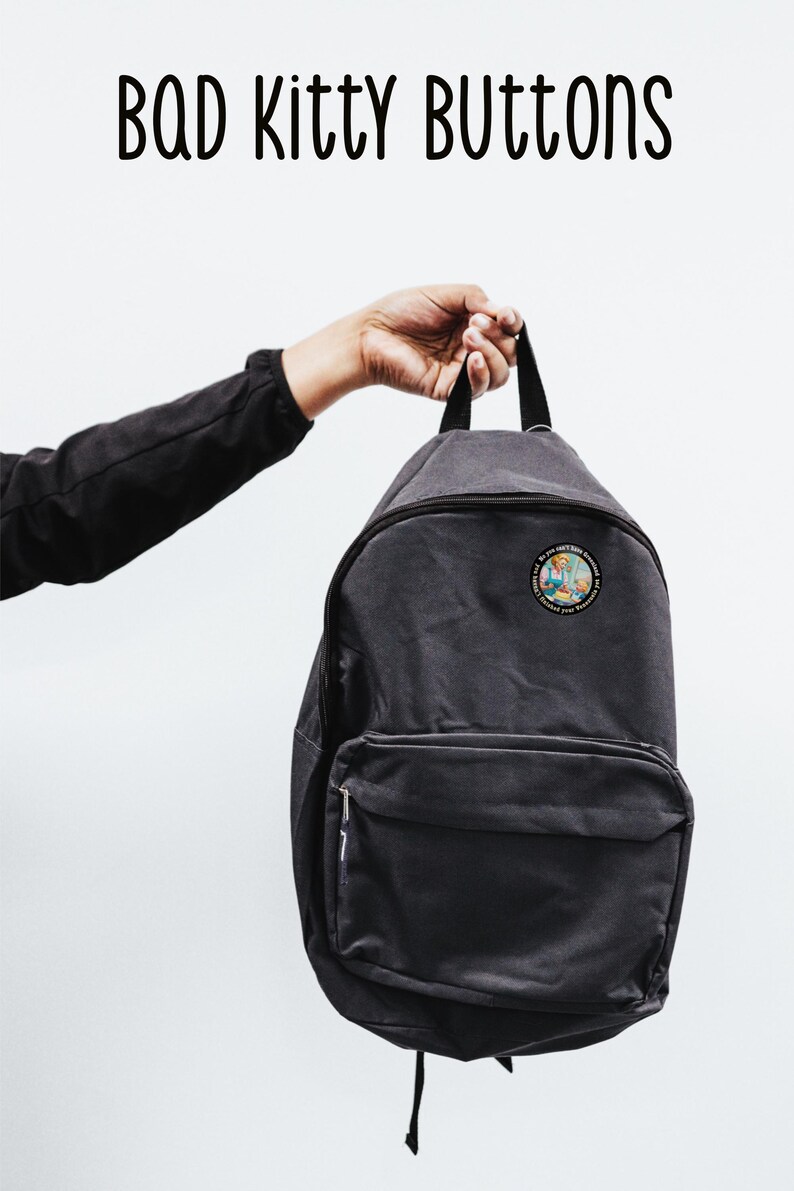 Op de afbeelding: Een donkergrijze rugzak met een patch met een cartoonafbeelding. De rugzak heeft een voorvak en een handvat aan de bovenkant. De tekst "BAD Kitty BUTTONS" staat boven de rugzak.