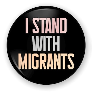 May include: Black button with the text "I STAND WITH MIGRANTS" in pink, gray, and orange lettering.