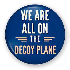 May include: A circular pinback button with a dark blue background. The text WE ARE ALL ON THE DECOY PLANE is printed in a light cream color in a sans-serif font. Two horizontal lines are positioned between the words ON and THE.