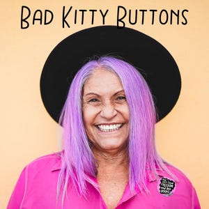 Puede incluir: Una persona sonriente con cabello morado y un sombrero negro, vistiendo una camisa rosa con un bot&oacute;n que dice "You can't Love Freedom and Ban Books". El texto "BAD KITTY BUTTONS" est&aacute; encima de la cabeza de la persona sobre un fondo amarillo.