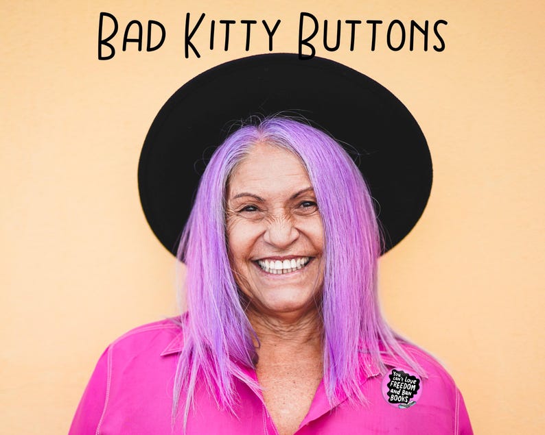 Puede incluir: Una persona sonriente con cabello morado y un sombrero negro, vistiendo una camisa rosa con un bot&oacute;n que dice "You can't Love Freedom and Ban Books". El texto "BAD KITTY BUTTONS" est&aacute; encima de la cabeza de la persona sobre un fondo amarillo.