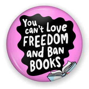 Puede incluir: Un bot&oacute;n redondo rosa con el texto blanco "You can't Love FREEDOM and Ban BOOKS" sobre un fondo negro ondulado. Una ilustraci&oacute;n de un libro abierto se encuentra en la parte inferior.