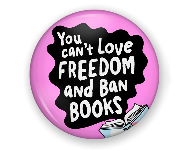 Puede incluir: Un bot&oacute;n redondo rosa con el texto blanco "You can't Love FREEDOM and Ban BOOKS" sobre un fondo negro ondulado. Una ilustraci&oacute;n de un libro abierto se encuentra en la parte inferior.